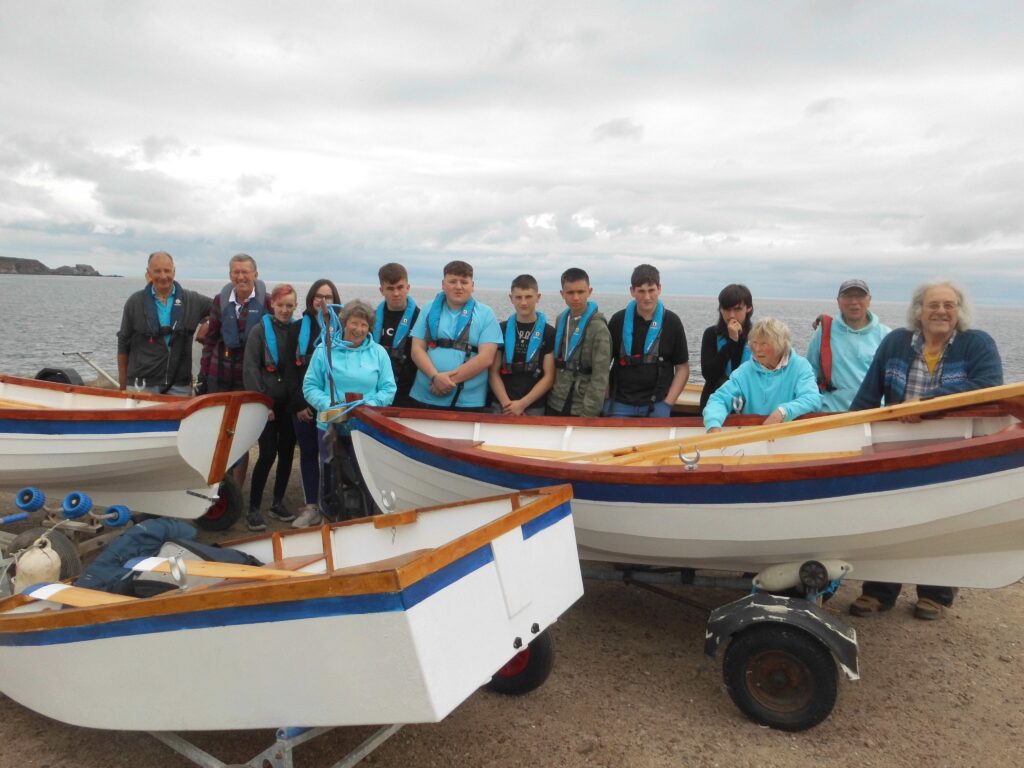 Image of Banff Academy Students with sea school boat builders and coastal rowers with the boats they have built outside the sea school with the sea beyond.